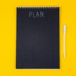 Σκούρο σημειωματάριο και λευκό στυλό επάνω σε κίτρινο γραφείο